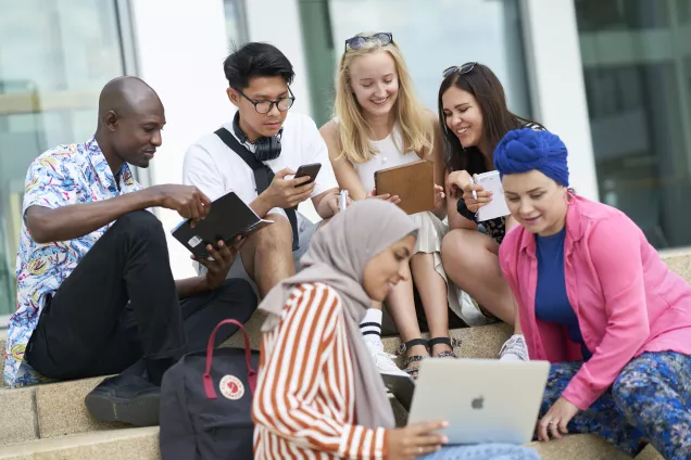 Foto på en grupp studenter som sitter utomhus i en trappa. De tittar på en laptop.