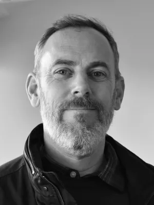 Black and white head shot profile photo of a man in grey beard with shirt, sweater and jacket.