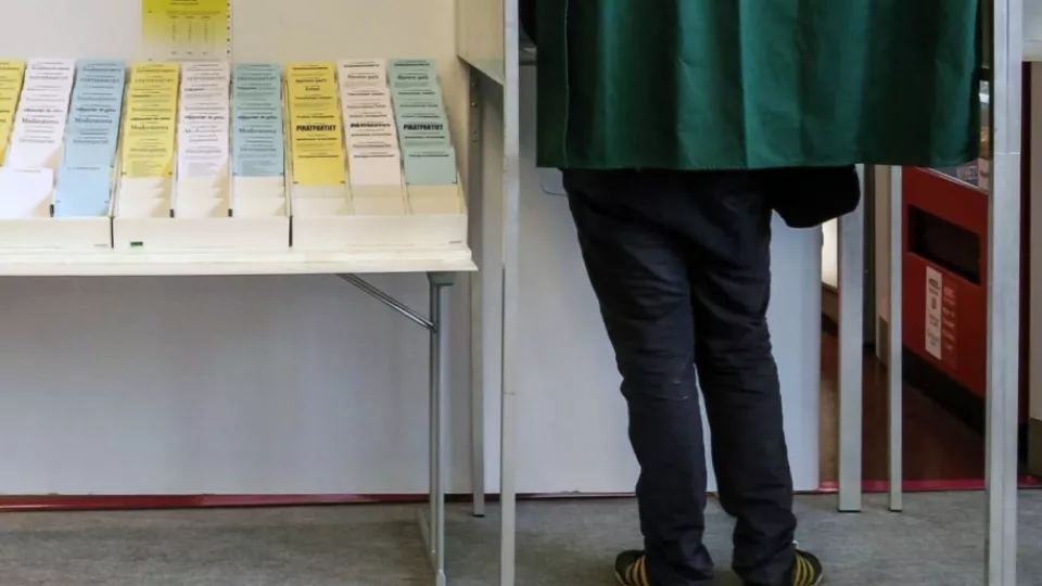 En vallokal där en person står bakom ett skynke och röstar. Bredvid står ett bord med valsedlar. 