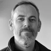 Svartvitt profilfoto av en man i grått kort skägg med mörk skjorta, tröja och jacka.